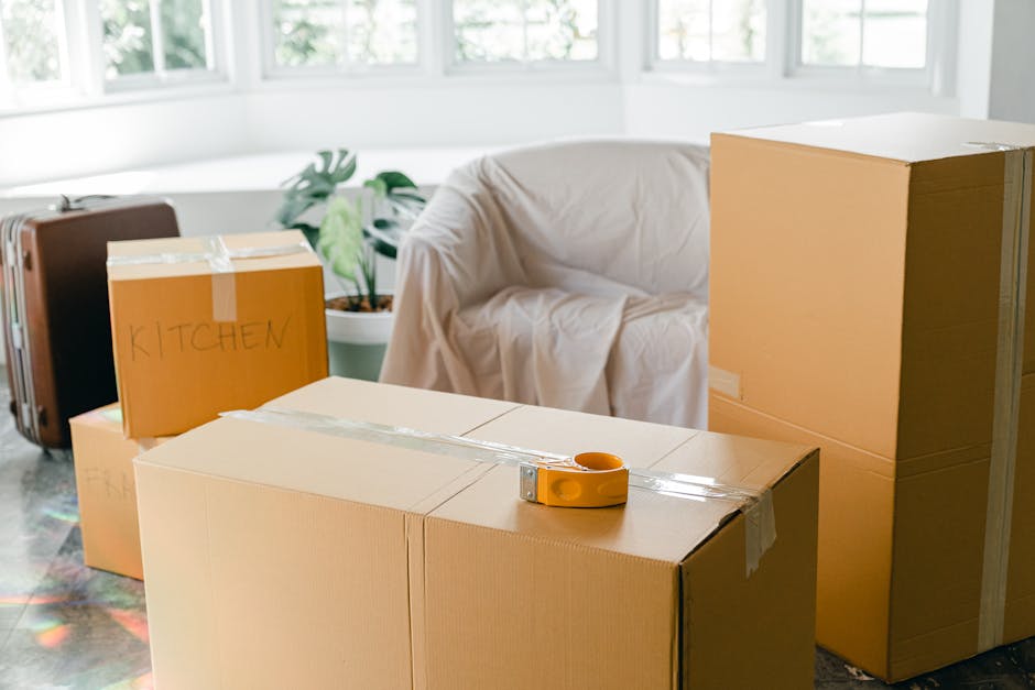 A man and a woman are involved in a home relocation process indoors, surrounded by numerous brown cardboard boxes of various sizes, some with black handles, which are stacked against the wall and scat