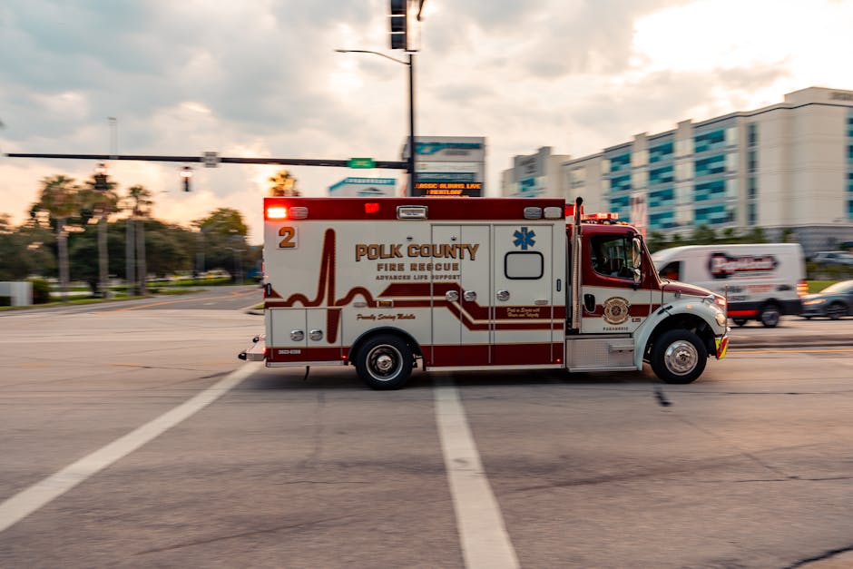 A red and white Polk County Fire Rescue ambulance is stationary at an intersection during daytime with urban buildings, trees, and traffic lights in the background. The vehicle is positioned parallel 