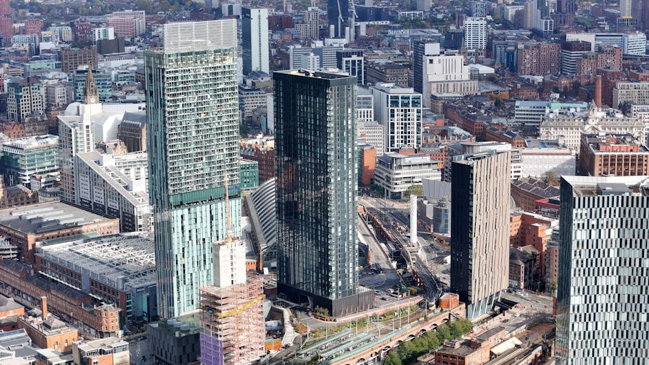 An aerial view of a major city showing a diverse array of high-rise and mid-rise office buildings, residential towers, and commercial structures. In the foreground, roads and parking lots are visible