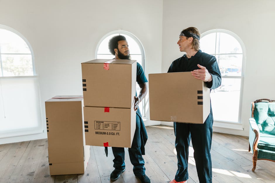 A male removals worker wearing a white t-shirt and black cap holds a cardboard box while a woman in a white cap and black sleeveless top writes on a clipboard, both standing inside a moving van filled